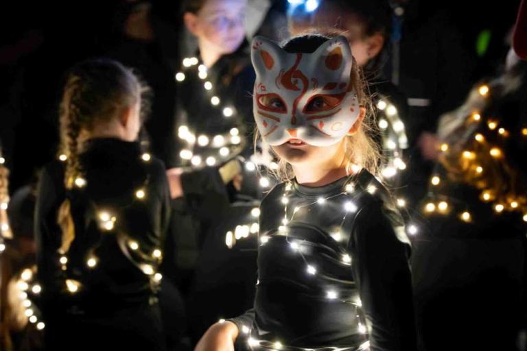 Image of a girl with a white and red mask dressed in black with fairy lights attached to her jumper