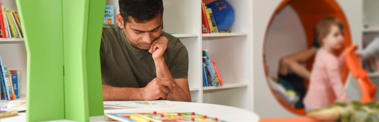 Man reading a book at the local library with a little girl in the background playing